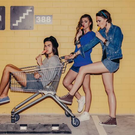 Two young young modern happy girls and handsome boy having fun in front of yellow brick wall. Beautiful ladies hold mall trolley with man inside. Everybody eat ice cream.の写真素材