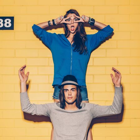 Young modern happy couple in love having fun in front of yellow brick wall. Beautiful longhaired girl in blue hoodie make mask with her fingers and her handsome husband rise his nads up. Boy sit on his skateboard.の写真素材