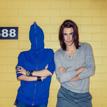 Young modern happy couple in love having fun in front of yellow brick wall. Beautiful lady in blue hoodie zipped all way up with her head and her handsome husband who looks to the camera. Both with crossed arms.の写真素材