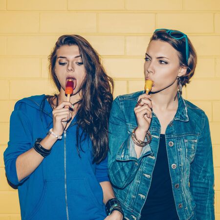 Lifestyle portrait of two beautiful best friends hipster girls wearing stylish bright outfits and having great time. Standing together near yellow brick wall enjoying day off, have fun and suck ice-cream.の写真素材