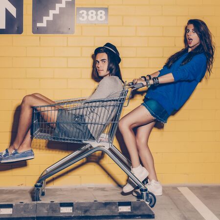 Young modern happy couple in love having fun in front of yellow brick wall. Beautiful lady in blue hoodie hold mall trolley with her handsome husband in hat. Both looks to the camera.の写真素材