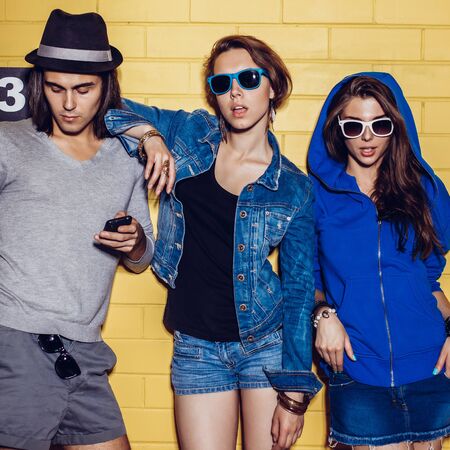 Lifestyle portrait of beautiful best friend hipsters wearing stylish bright outfits and sunglasses and having great time. Girls are looking to the camera in front of yellow brick wall enjoying day off and have fun. Boy with smartphone cellphone.の写真素材
