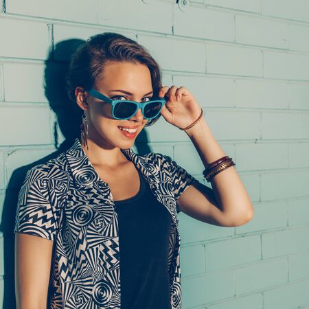 Young beautiful sexy lady in modern jacket and black t-shirt playing with her sunglasses while smiling to the camera in front of blue brick wall.の写真素材