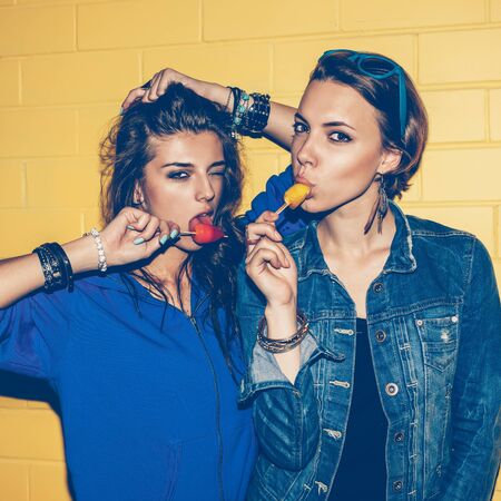 Lifestyle portrait of two beautiful best friend hipster lady wearing stylish bright outfits and having great time. Standing together near yellow brick wall enjoying day off, have fun and eating sweet cold ice-cream.の写真素材