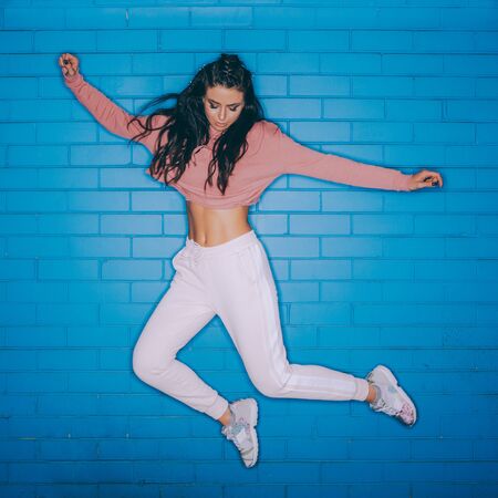 Fashion studio flash portrait of athletic young cute woman in vogue sport suit jump against blue brick wall. Urban style, healthy girl lifestyle and sport concept. Hipster city life.の写真素材