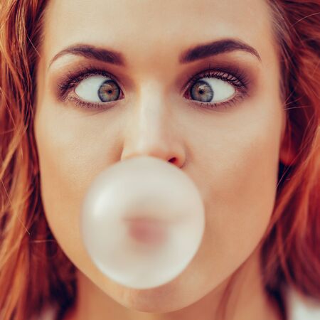 Funny portrait of beautiful young cute woman in white vogue suit look to her nose while blowing gum bubble against red wall. Urban style girl, quick lifestyle and work concept. Hipster city life.の写真素材