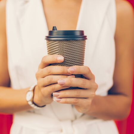 Close up of blank white to go cup of hot tea or coffee in hands of young business woman in white vogue suit against red wall. Urban style girl, quick lifestyle and work concept. Hipster city life.の写真素材