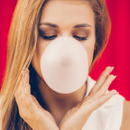 Funny portrait of beautiful young cute woman in white vogue suit look blowing gum bubble against industrial red wall. Urban style girl, quick lifestyle and work concept. Hipster city life.の写真素材