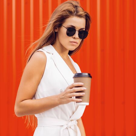 Fashion portrait of beautiful young business woman in white vogue suit with to go cup of hot tea or coffee against red wall. Urban style girl, quick lifestyle and work concept. Hipster city life.の写真素材