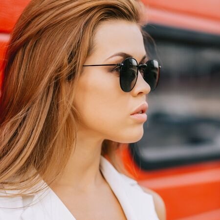 Fashion portrait of beautiful young business woman in white vogue suit and black sunglasses against industrial red wall. Urban style girl, quick lifestyle and work concept. Hipster city life.の写真素材