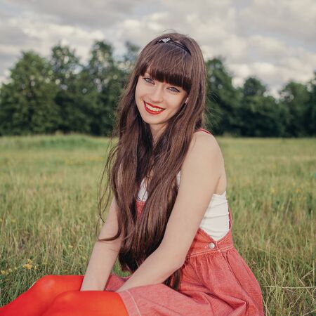 Pretty girl in red sarafan on the green grass. A beautiful girl with long hair coquettishly smiling. Young modern city woman look to the camera. Urban lifestyle holidays. Farm life.の写真素材