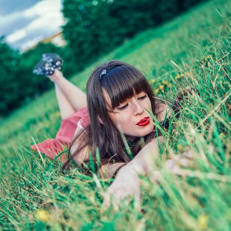 Pretty girl in red sarafan on the green grass. A beautiful girl with long hair coquettishly smiling. Young modern city woman look to the camera. Urban lifestyle holidays. Farm life.の写真素材