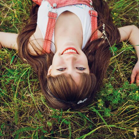 Pretty girl in red sarafan on the green grass. A beautiful girl with long hair coquettishly smiling. Young modern city woman look to the camera. Urban lifestyle holidays. Farm life.の写真素材