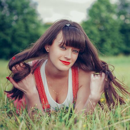 Pretty girl in red sarafan on the green grass. A beautiful girl with long hair coquettishly smiling. Young modern city woman look to the camera. Urban lifestyle holidays. Farm life.の写真素材