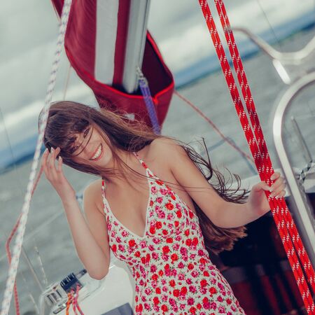 Beautiful girl in light dress standing on the deck of sailboat. Beautiful girl with long hair coquettishly smiling. Young modern city woman look to the camera. Urban lifestyle holidays. Farm life.の写真素材