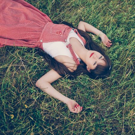 Pretty girl in red sarafan on the green grass. A beautiful girl with long hair coquettishly smiling. Young modern city woman look to the camera. Urban lifestyle holidays. Farm life.の写真素材