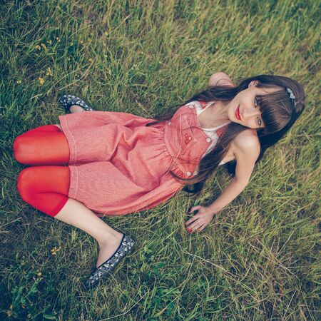 Pretty girl in red sarafan on the green grass. A beautiful girl with long hair coquettishly smiling. Young modern city woman look to the camera. Urban lifestyle holidays. Farm life.の写真素材