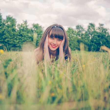 Pretty girl in red sarafan on the green grass. A beautiful girl with long hair coquettishly smiling. Young modern city woman look to the camera. Urban lifestyle holidays. Farm life.の写真素材