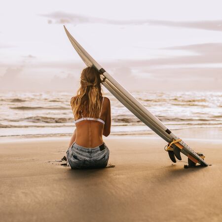 Young surfer woman in sexy white bikini with blank surfing shortboard sit on sand at sunset ocean. Modern family lifestyle, people water sport adventure camp and extreme swim on summer vacation.の写真素材