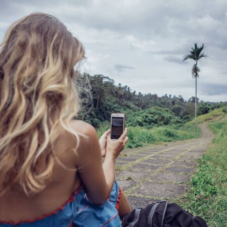 Beautiful young woman in blue dress seat at stone hill path in Asia and take pictures on her smartphone. Girl travel and explore world. Long lonely road in mountains. Travel and adventure mockup.の写真素材