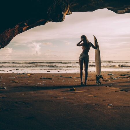 Young surfer lady with surf board pose in cave at beach. Garbage and plastic on sand. Concept of ecological problem and disaster. People water sport adventure camp and extreme swim on summer vacationの写真素材