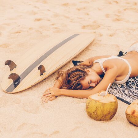 Happy girl lonely rest on a sand beach near wooden surf surfboard board at sunrise or sunset. Woman lies near green coconut with fresh coco water. Vacation concept. Summer holidays. Tourism, sport.の写真素材