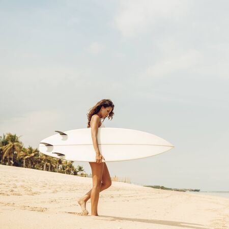Beautiful fit surfing girl in sexy bikini swimwear with white surf shortboard surfboard blank board at sunrise or sunset on sand beach walk to ocean. Vacation concept. Summer holidays. Tourism, sport.の写真素材