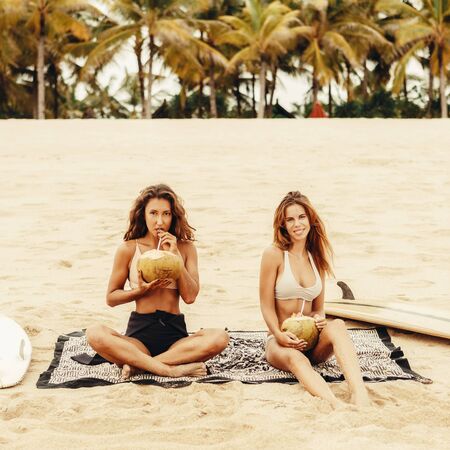 Two beautiful young girl in sexy bikini drink fresh coconut cool coco water on a sand beach after surfing with wooden surf white surfboard board at sunrise sunset. Summer holidays. Tourism, sport.の写真素材