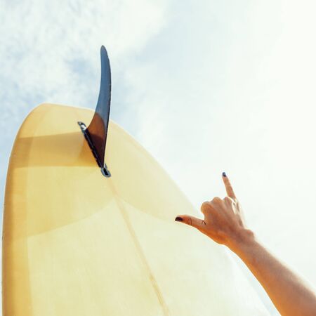 Close up of a woman hand show surfing sign shaka in front of yellow single fin surf longboard surfboard board on a sand beach at sunset or sunrise. Modern active sport lifestyle and summer vacation.の写真素材