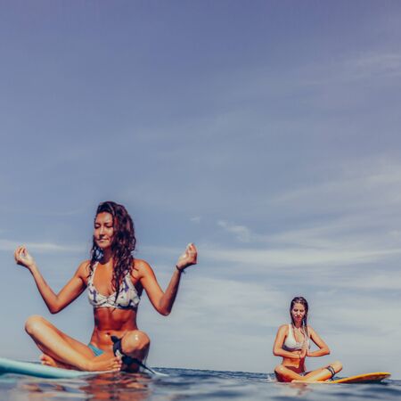 Two beautiful sexy surfing girl doing yoga and balancing into the ocean on a big longboard surf surfboard board on sunrise or sunset. Modern active sport lifestyle and summer vacation.の写真素材