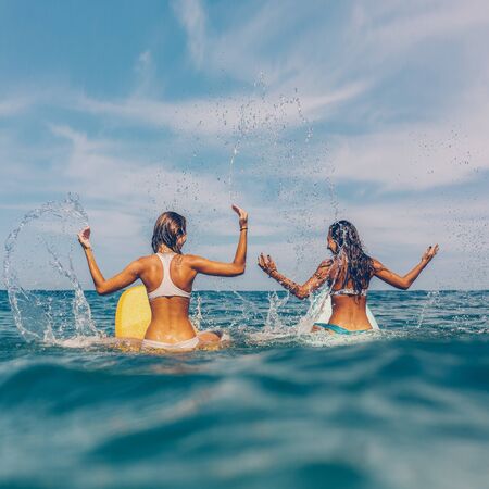 Two beautiful sporty surfing girl in sexy bikini sit on a big longboard surf surfboard board and make splashes on sunrise or sunset in the ocean. Modern active sport lifestyle and summer vacation.の写真素材