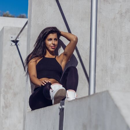 Beautiful young girl with long legs in black sexy leggings with pretty athlete muscular body rest after yoga. Crossfit training urban area street gym city exercise routine healthy lifestyle.の写真素材