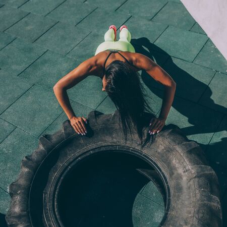 Top view of young woman in bright sexy leggings with pretty athlete muscular body doing push ups exercise on big tire. Crossfit training urban area street gym city workout routine healthy lifestyle.の写真素材
