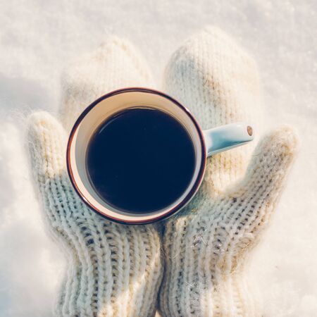 Young beautiful girl in hipster white knitted mittens drink hot tea on a bright winter day. Close-up of a cup with hot liquid in hand. Trees in pure first snow on a background. Top view.の写真素材