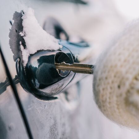 Young beautiful woman in hipster white knitted mittens try to open vehicle on a winter morning. Close-up of a key inserted into the lock of frozen car door. Transportation and ownership concept.の写真素材