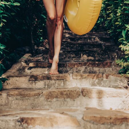 Close up of a sexy young lady in striped bikini and white t-shirt with yellow inflatable swimming ring walking on stone steps in a tropical garden. Outdoor lifestyle picture on a sunny summer day.の写真素材