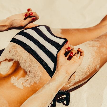 Sporty young beautiful lady correct her striped sexy bikini while sunbathing with sandy fit butt on the tropical ocean shore background. Outdoor lifestyle picture on a hot sunny summer day.の写真素材