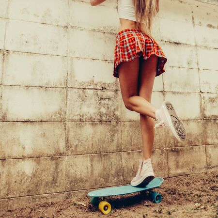 Sporty young woman with fit butt in a red tartan mini skirt stands on blue penny skateboard trying to look over the fence of a tropical garden. Outdoor lifestyle picture on a sunny summer day.の写真素材