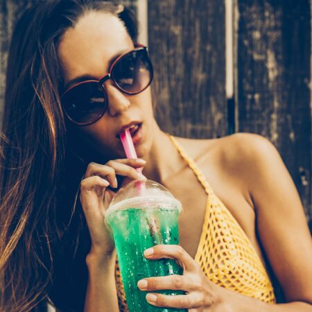 Sporty young girl in a yellow knitted swimsuit trying a green refreshing drink in front of wooden fence in a tropical garden. Outdoor lifestyle picture on a sunny summer day.の写真素材