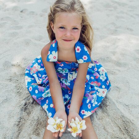 Outdoor portrait of cute small blonde girl in floral dress sitting on the beach sand. Little lady hold frangipani tropical flowers and smiles to the camera. Summer sunny day. Happy Mothers day.の写真素材