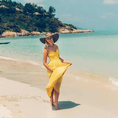 Carefree beautiful fashion blonde female in beach straw hat holding her long yellow dress and walking barefoot by sand of tropical beach. Natural woman beauty. Lady looking to the camera and dance.の写真素材