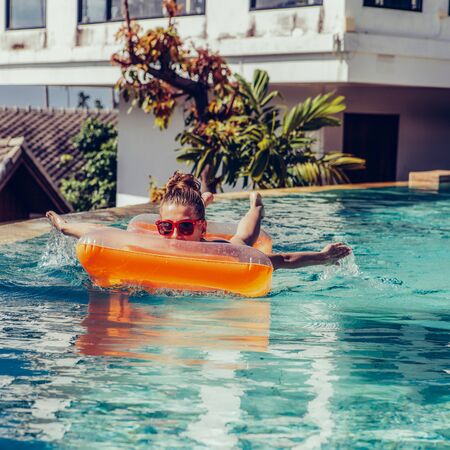 Happy beautiful female in trendy swimsuit and sunglasses have fun at hotel swimming pool party. Young sexy girl enjoying holiday, having fun and win swimming race on inflatable orange mattress.の写真素材