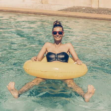 Amazing beautiful woman in swimsuit and sunglasses have fun at swimming pool party. Portrait of sexy girl enjoying hot day, having fun with inflatable yellow ring in water and show her feet and smile.の写真素材