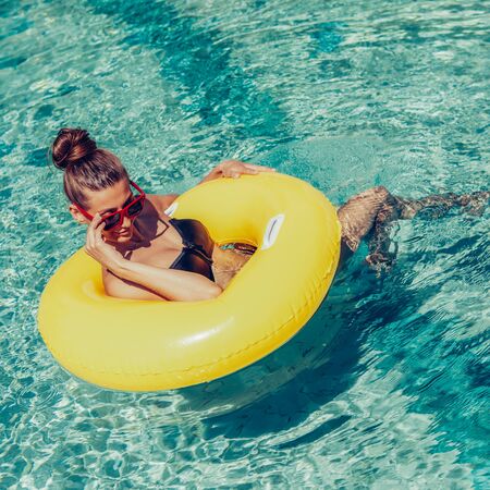 Amazing beautiful lady in black swimsuit correct trendy sunglasses and have fun at swimming pool party. Outdoor portrait of sexy girl enjoying hot summer day, swim with inflatable yellow ring.の写真素材