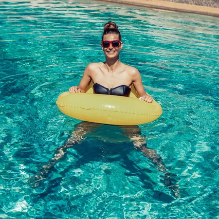 Amazing beautiful woman in swimsuit and sunglasses have fun at swimming pool party. Outdoor portrait of sexy girl enjoying hot day, having fun with inflatable yellow ring in water and smile to camera.の写真素材