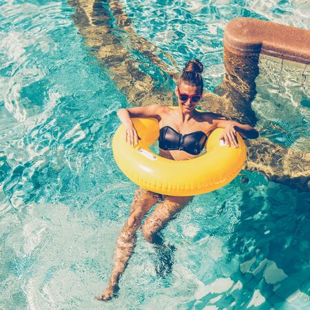 Happy beautiful woman in trendy swimsuit and sunglasses have fun in hotel swimming pool party. Young sexy girl enjoying hot day, having fun and sunbathing on inflatable yellow ring in water.の写真素材