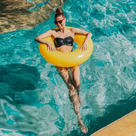 Happy beautiful lady in trendy swimsuit and sunglasses have fun in hotel swimming pool party. Young sexy girl enjoying holiday, having fun and sunbathing on inflatable yellow ring in cool water.の写真素材