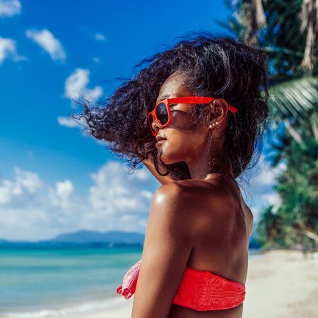 Outdoor lifestyle portrait of black beautiful teenage girl in pink bra and sunglasses. Young hipster woman play with her hair. She is happy. Sunny hot summer day at tropical ocean beach with palms.の写真素材