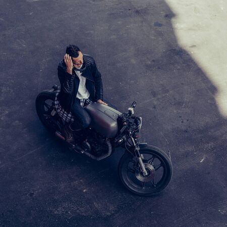 Top view of a handsome rider guy in black biker jacket and checkered shirt correct hes hair sit on classic style cafe racer motorcycle. Bike custom made in vintage garage. Brutal fun urban lifestyle.の写真素材