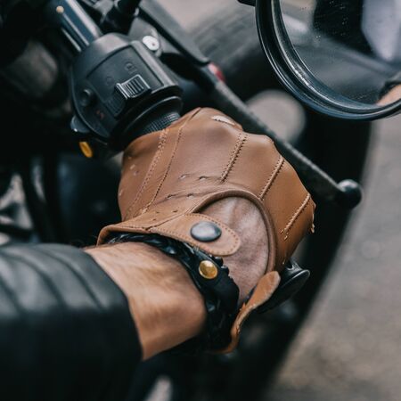 Closeup of a hipster biker man hand in leather glove hold throttle control of classic style cafe racer motorcycle. Bike custom made in vintage garage. Brutal fun urban lifestyle. Outdoor portrait.の写真素材